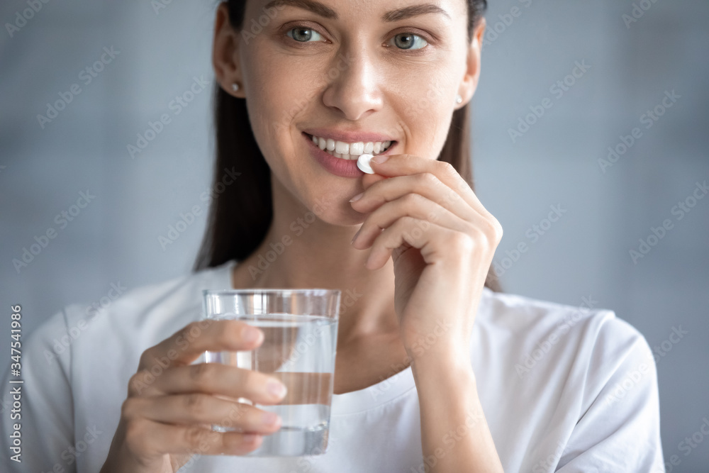 Close up image young adult beautiful 35s woman smiles takes pill holding glass of water, concept of minerals and vitamins for female, hair skin nail care dietary supplement, daily dosage of medication - obrazy, fototapety, plakaty 