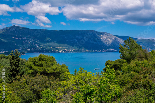 Beautiful summer landscape – calm sea water surface, white yacht, green trees and mountains on the horizon. Corfu Island, Greece. © elenakirey