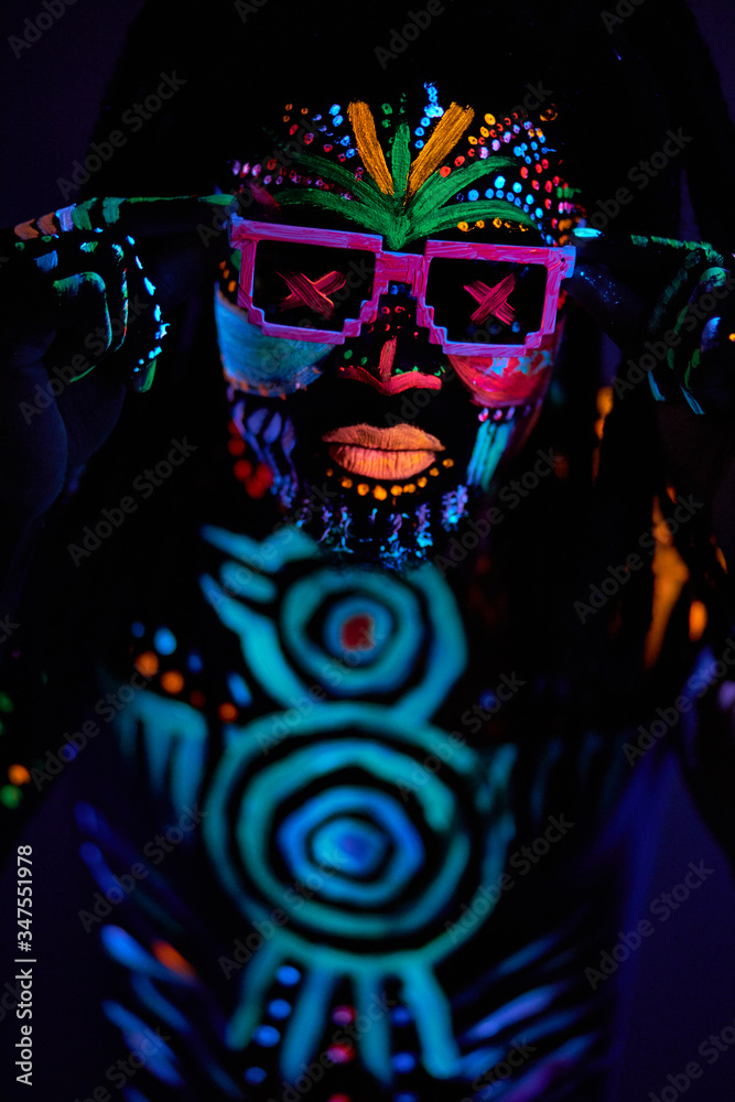 portrait of cool african man with fluorescent body art dancing isolated in dark space. dj man perform dance, mysterious dancer with colorful prints on face and body
