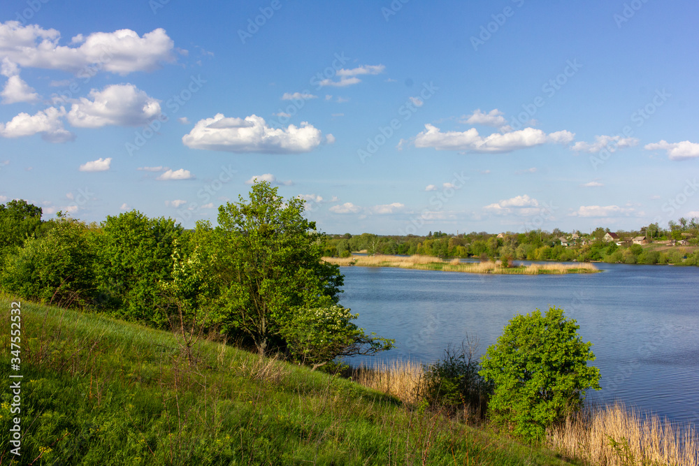 Idyllic summer landscape with river, hill, green trees, blue sky, and white clouds. Natural warm light