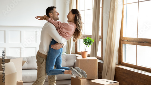 Happy young couple celebrating moving day, hugging in new apartment, loving husband lifting smiling wife, standing in living room with cardboard boxes with belongings, relocation and mortgage photo