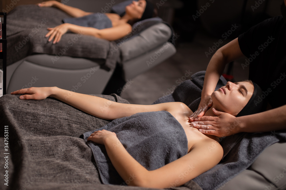 two young caucasian ladies get anti aging massage on neck, attractive women have rest at spa wellbeing salon, skin care, face lifting concept