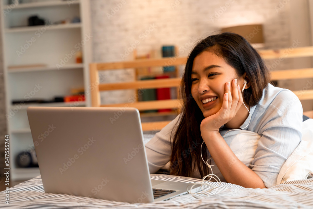 Fototapeta premium Young smiling Asian woman in casual clothing and lying on bed with laptop computer. Female freelancer working from home on notebook