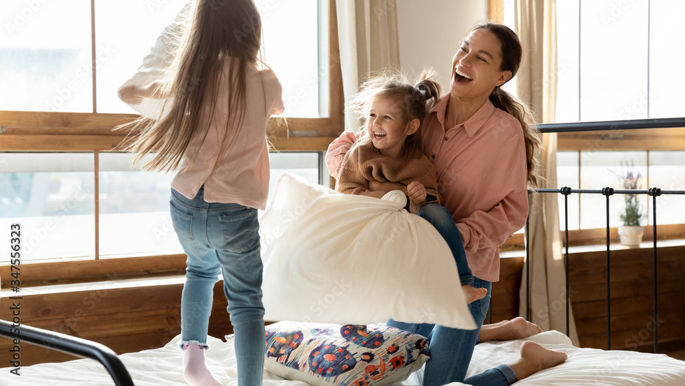 Fototapeta premium Happy adorable little daughters with young mother pillow fighting in bedroom, laughing and jumping, two cute preschool girls having fun with mum, playing active funny game at home