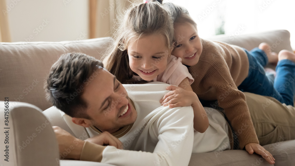 Happy father with two little daughters relaxing on couch close up, smiling young dad and preschool cute girls hugging, lying on cozy sofa in living room, family having fun on weekend