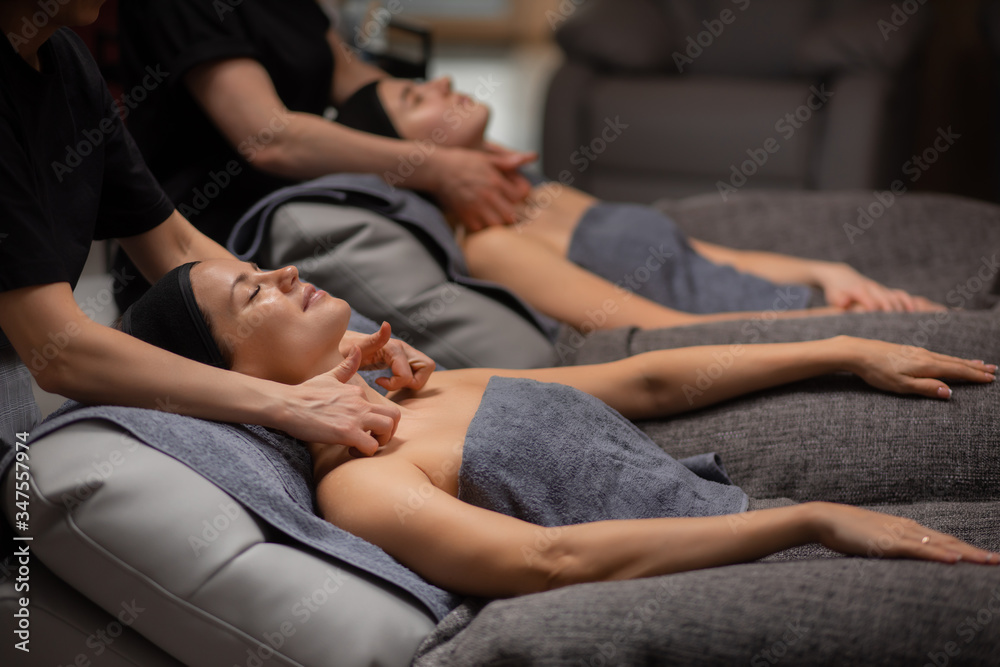 two young caucasian ladies get anti aging massage on neck, attractive women have rest at spa wellbeing salon, skin care, face lifting concept
