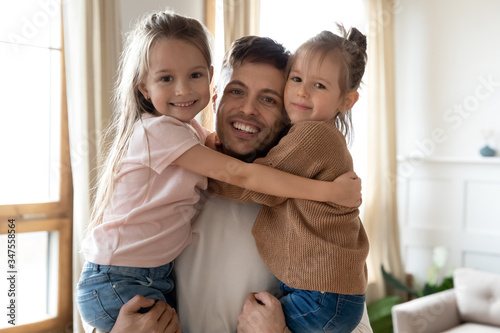 Head shot portrait smiling excited father holding little daughters in arms, looking at camera, happy young dad hugging two cute preschool girls, posing for family photo together at home