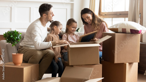 Happy parents with children unpacking cardboard boxes with belongings together, sitting on couch in new apartment, smiling mother, father and two daughters looking at family portrait in photo frame