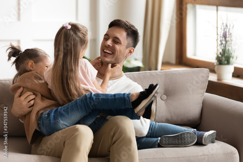 Happy father playing with two little daughters, tickling, sitting on couch at home, cute preschool girls sitting on dad laps, playing funny game, family spending free time together