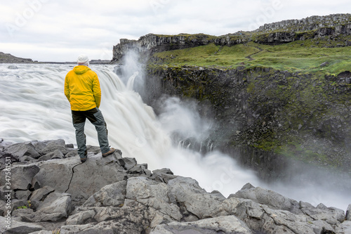 A man with yellow jacket stands on the edge looking towards the powerful dettifoss waterfall in northern iceland during early morning soft light. Traveler and holiday concept.