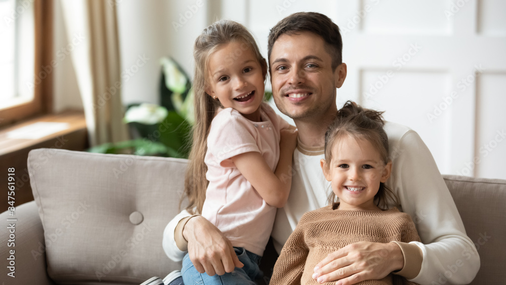 Head shot portrait happy father hugging two little daughters, sitting on couch at home, smiling young dad with preschool cute girls looking at camera, posing for family photo together Head shot portrait happy father hugging two little daughters, sitting on couch at home, smiling young dad with preschool cute girls looking at camera, posing for family photo together