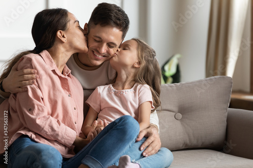 Mother and little daughter kissing father on cheek, sitting on cozy sofa, happy family enjoying tender moment at home, mum and child expressing love, congratulating with birthday or fathers day
