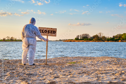 Man wearing protective suit stands with warning sign in hands with inscription closed along the sand beach of the river. Contaminated water, quarantine, virus outbreak, crime scene concept