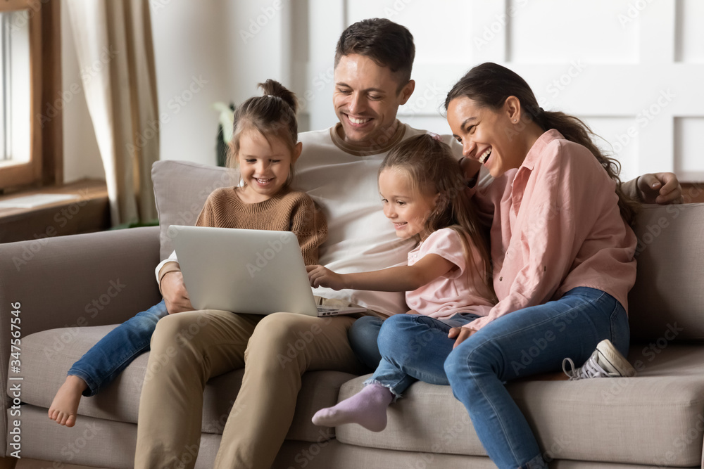 Happy family with two little daughters using laptop together, sitting on cozy sofa at home, smiling father and mother with preschool girls sisters looking at computer screen, shopping online Happy family with two little daughters using laptop together, sitting on cozy sofa at home, smiling father and mother with preschool girls sisters looking at computer screen, shopping online