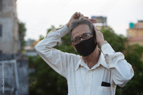 portrait of an Indian old man with corona preventive mask combing his hair on a rooftop during sunset in home isolation.Indian lifestyle, disease and home quarantine.