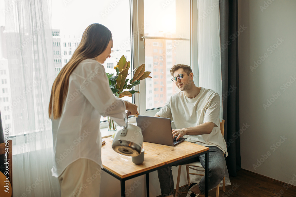 kind caucasian woman treats her husband with tasty tea while he is working from home, using laptop. merciful wife loves husband, support him. freelance, relationships, business at home
