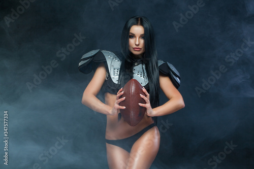 American football. Young sporty brunette wearing sexy uniform of rugby football player posing with ball in smoke isolated on black background
