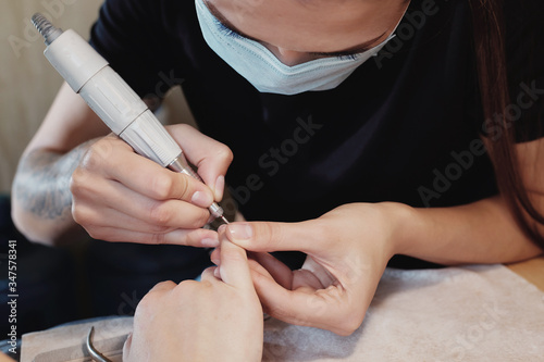 The manicure master is doing nails to the client in a medical mask with drill. close-up