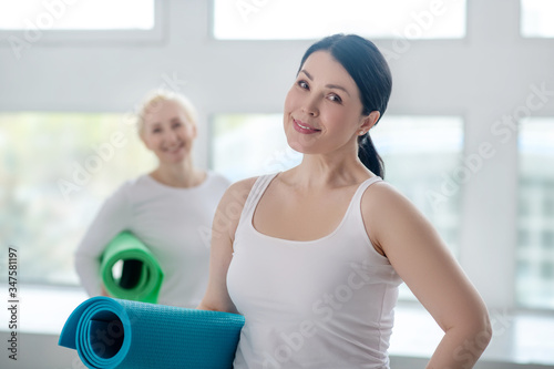 Tableau sur toile Brunette female holding yoga rug and smiling, blonde female standing in the back