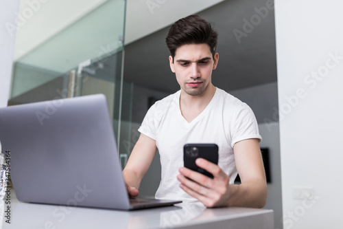 Attractive young man working from home sitting at a table kitchen as he reads a text message on his mobile