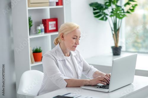 Blonde middle-aged practitioner sitting at her desk and talking to patient online