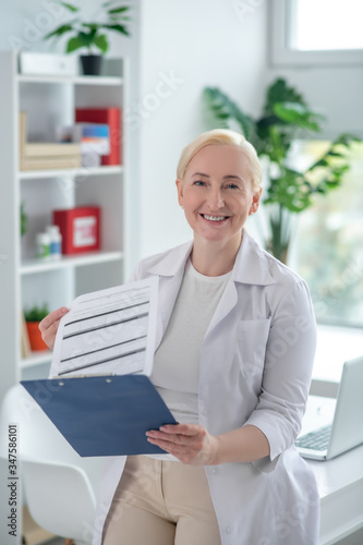 Blonde middle-aged doctor reading medcial history and looking positively photo