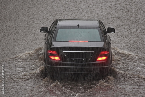 Black passenger car is driving through a large puddle on road during heavy rain, back view, meteorological cataclysm