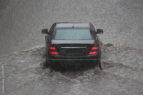 One black car is driving through a big puddle with wave on road during heavy rain, local inundation, rear view