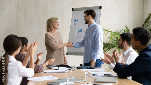 Happy young businessman congratulation hand shaking 60 year old businesswoman after success flipchart presentation new project in boardroom at meeting. Manager shaking hand mature good employee.