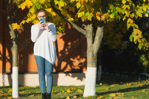 Millennial woman texting on her blue smartphone. Full length outdoor portrait of woman in spectacles wearing white sweater and jeans on warm October evening.