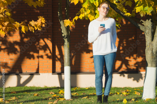 Millennial woman smiling shrewdly at camera, holding her smartphone in right hand. Full length outdoor portrait of woman in spectacles wearing white sweater and jeans on joyful October evening.