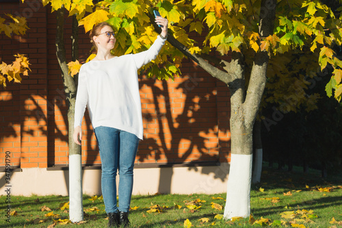 Millennial woman video chatting on tranquil day outside. Full length portrait of caucasian woman smiling at phone, wearing spectacles, baggy white sweater and plain jeans on joyful October evening.