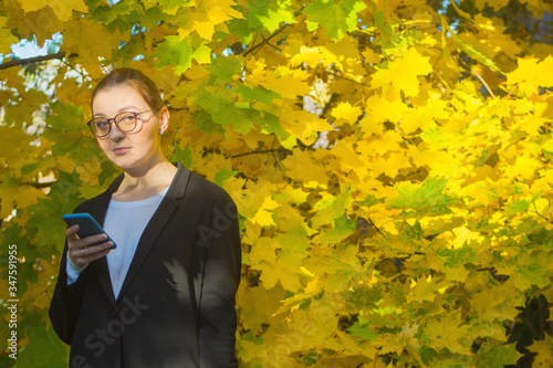 A woman is looking slightly surprised at the camera while holding her smartphone at chest level on a warm autumn day. Calm single person in tranquil outdoor environment.