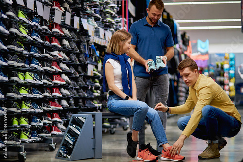 affable caucasian consultant man of sportswear store offer beautiful sportive sneakers to woman customer came in shop with boyfriend, she sit trying on