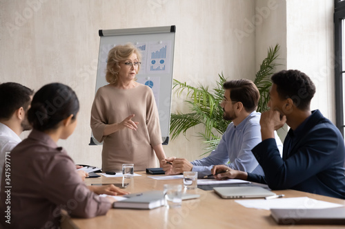 Confident mature 60 year old businesswoman presentation new project in boardroom at company meeting. Adult woman coach auditor speaks about business on flip chart graphs background. photo