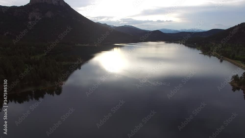 Aerial view of reflection of sun on lake amidst mountains, drone moving forward over water near forest against sky during sunset - Lake Wellington, Colorado Aerial view of reflection of sun on lake amidst mountains, drone moving forward over water near forest against sky during sunset - Lake Wellington, Colorado