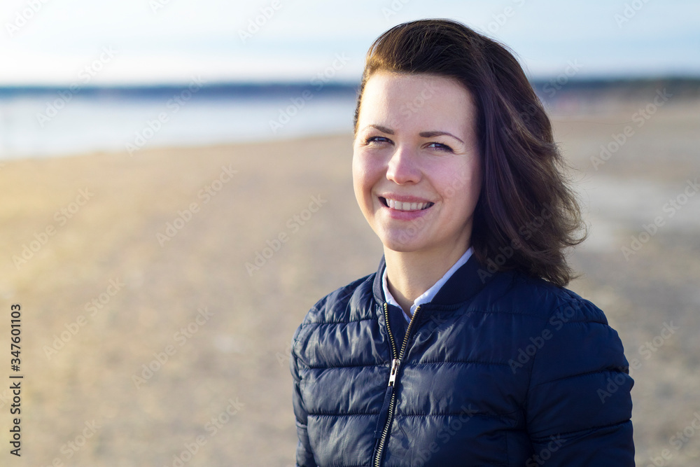 Portrait of a beautiful positive brunette girl, young happy cheerful attractive woman having fun, smiling, looking at camera outdoors at beach, bay in cold weather, sunny day. Copy space. Portrait of a beautiful positive brunette girl, young happy cheerful attractive woman having fun, smiling, looking at camera outdoors at beach, bay in cold weather, sunny day. Copy space.