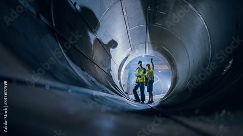 Two Heavy Industry Engineers Walking Inside Pipe, Use Laptop, Have Discussion, Checking Design. Construction of the Oil, Natural Gas and Biofuels Transport Pipeline. Industrial Manufacturing Factory