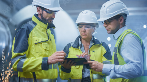 Three Heavy Industry Engineers Stand in Pipe Manufacturing Factory, Use Digital Tablet Computer, Have Discussion. Large Pipe Assembled. Design and Construction of Oil, Gas and Fuels Transport Pipeline