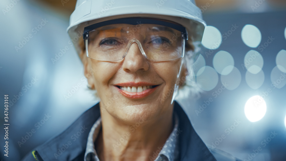 Portrait of Professional Heavy Industry Female Engineer Wearing Safety Uniform and Hard Hat, Glasses and Smiling Charmingly. In the Background Unfocused Industrial Factory where Welding Sparks Flying