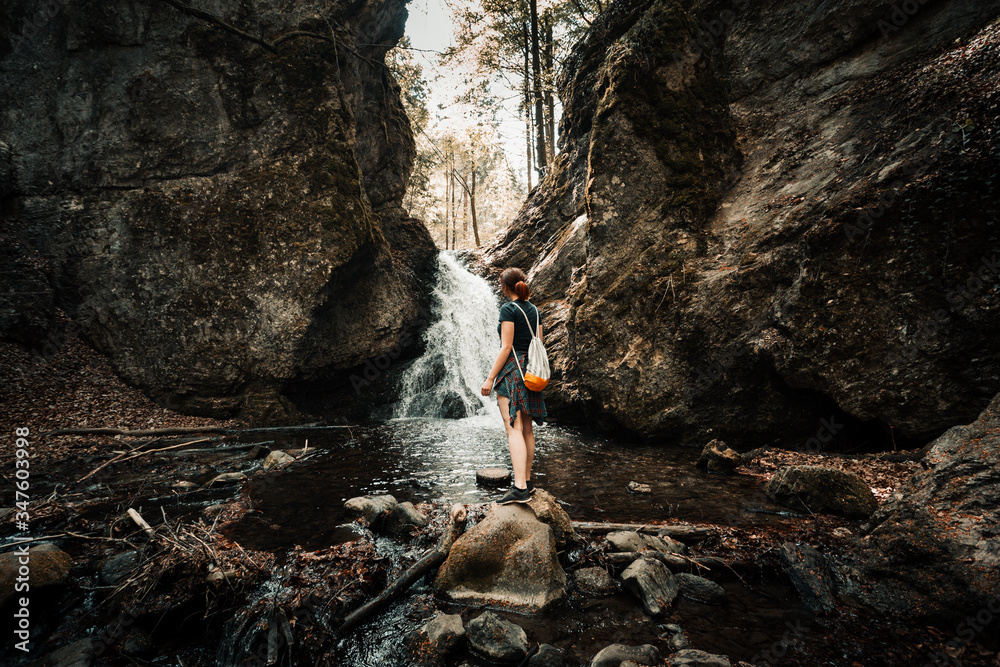 Woman (traveler and adventure) standing under the waterfall in dark and moody forest - surounded by stones and rocks. Folk tone shot of woman looking on the waterfall with backpack and relaxing. Woman (traveler and adventure) standing under the waterfall in dark and moody forest - surounded by stones and rocks. Folk tone shot of woman looking on the waterfall with backpack and relaxing.
