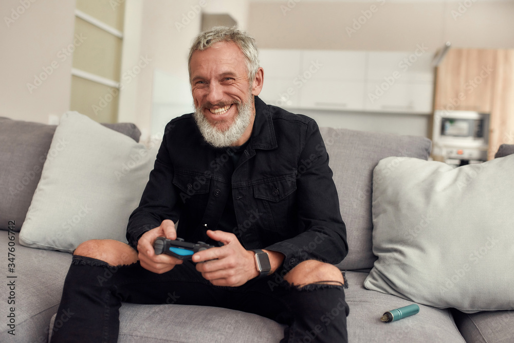 Relax. Happy bearded middle-aged man holding controller, playing video games, sitting on the couch at home. Weed vaporizer, dry herb vape pen lying next to him. Cannabis and weed legalization concept Relax. Happy bearded middle-aged man holding controller, playing video games, sitting on the couch at home. Weed vaporizer, dry herb vape pen lying next to him. Cannabis and weed legalization concept