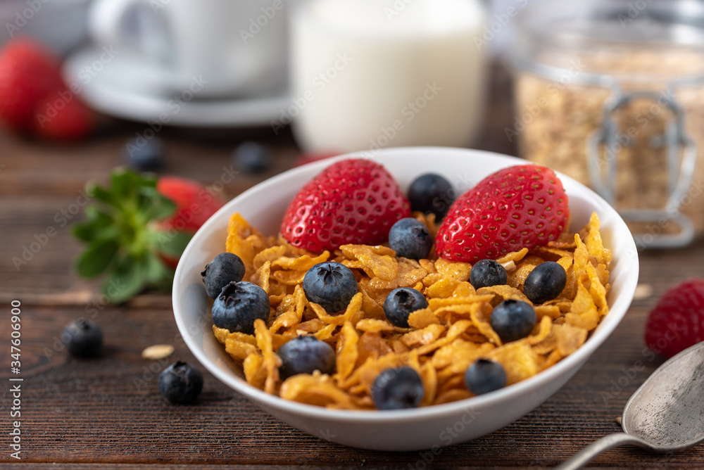 Cornflakes with blueberries and raspberries in white bowl on dark wooden desk.