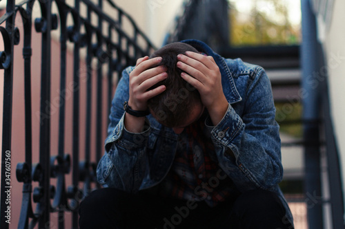 Sad young boy in jeans jacket with his hands on the head is sitting on city stair. photo