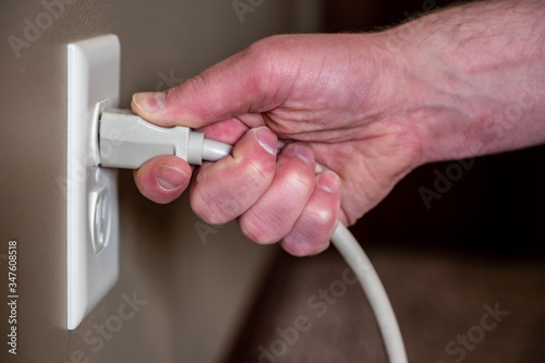 Caucasian hand gripping a three pronged power cord to remove it from a white outlet
