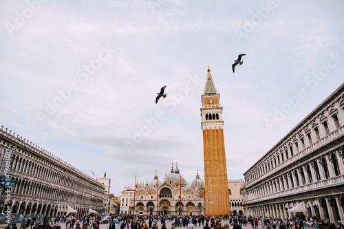 A huge bell tower made of red brick in Piazza San Marco - Campanile of St. Mark's Cathedral in Venice, Italy. Two seagulls flying by. A crowd of tourists at the foot of the tower in the square.