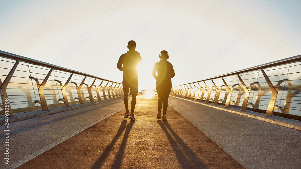 Perfect morning for run. Back view of young sporty couple jogging together on the bridge in the early morning at sunrise. Full length. Warm sunlight