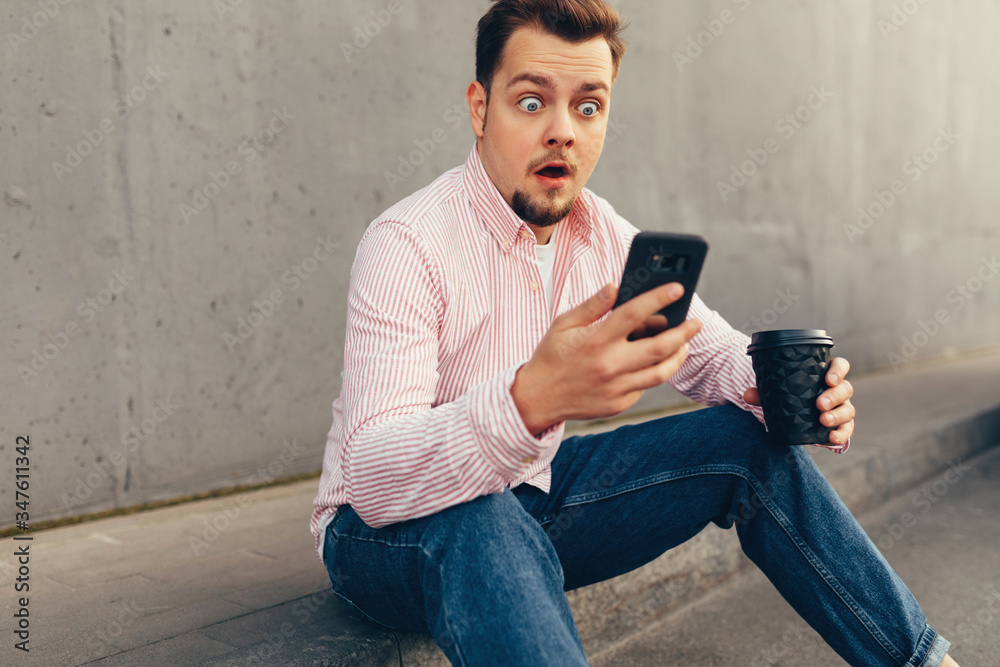 A young caucasian surprised man in striped shirt and blue jeans communicating in video chat or making selfie, sitting on the street curb with cup of coffee A young caucasian surprised man in striped shirt and blue jeans communicating in video chat or making selfie, sitting on the street curb with cup of coffee