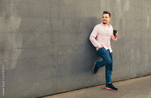 A young handsome stylish smiling guy standing at the gray wall and drinking coffee or tea from disposable cup outdoors. City lifestyle concept