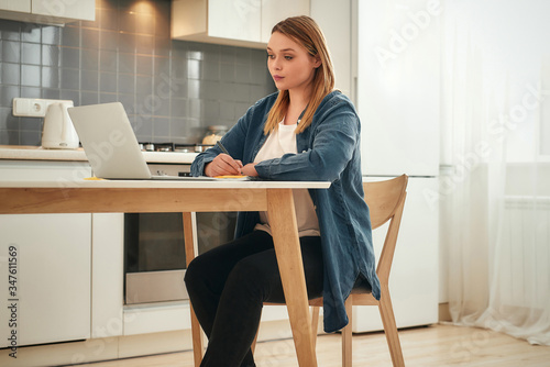 Concentrated at work. Young caucasian girl in casual clothes working from home, she is sitting at kitchen table at home and using laptop